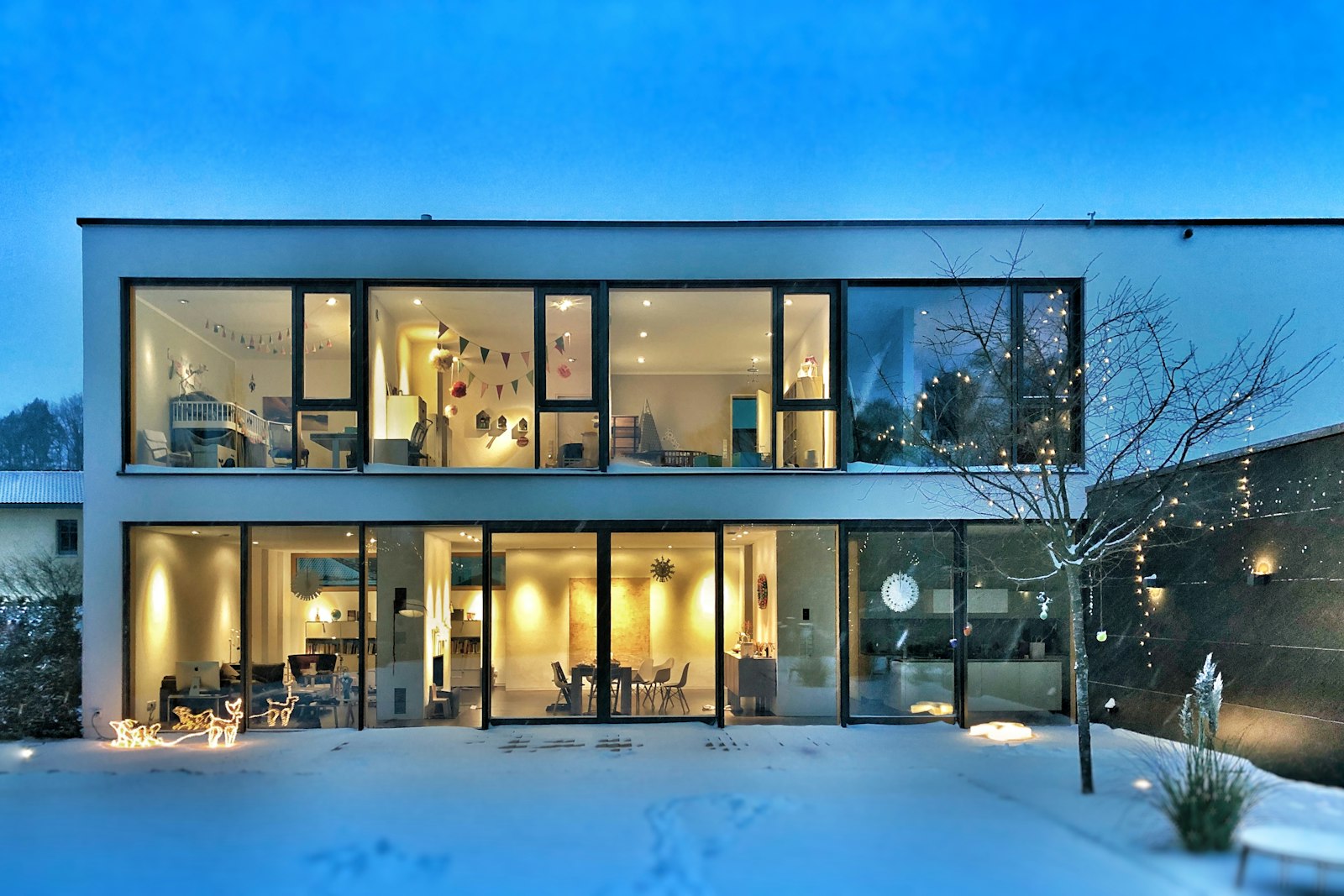 Maison contemporaine vitrée, jardin enneigé et lumières intérieures au crépuscule